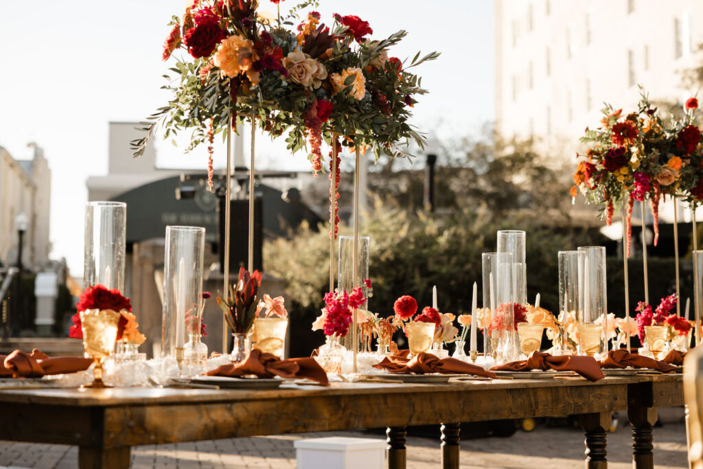 Maroon, Purple, and Orange Roses, Stock, Hypernicum Berry, Wisteria, Garden Rose, and Greenery Tall Centerpiece Ideas | Taper Candles with Hurricane Tube Glass | Dark and Moody Winter Wedding Reception Inspiration | Wooden Feasting Tables | Vintage Goblets | Lakeland Kate Ryan Event Rentals