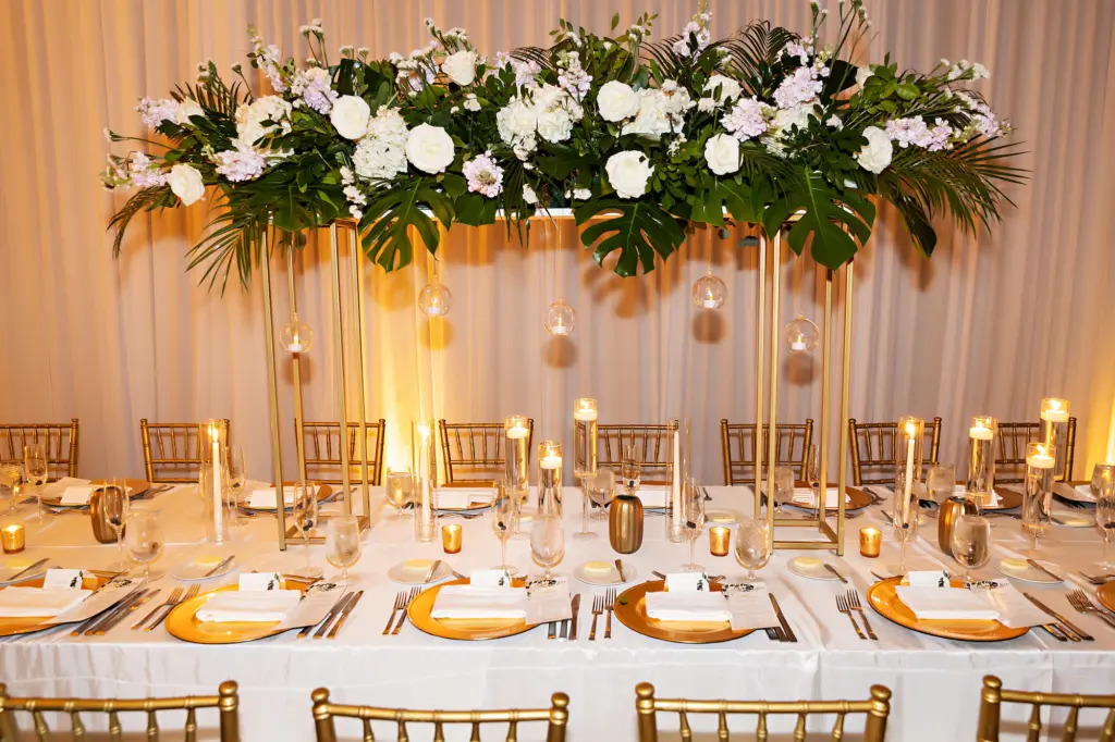 Long Feasting Sweetheart Table | Classic White and Gold Wedding Reception Inspiration with Neon Sign | Tall Flower Stands with Monstera, Palms, White Roses and Hydrangeas