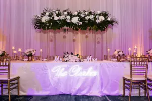 Long Feasting Sweetheart Table with Neon Sign | Classic White and Gold Wedding Reception Inspiration with Neon Sign | Tall Flower Stands with Monstera, Palms, White Roses and Hydrangeas