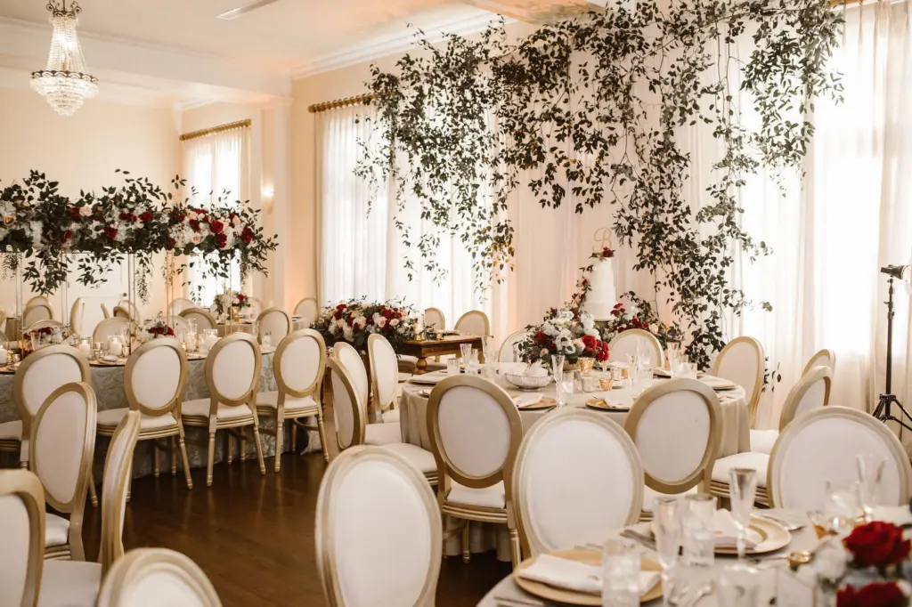 Tall Gold Flower Stand with Cascading Greenery, Burgundy, White, and Blush Rose Centerpiece Inspiration | Gold Chargers and Long Feasting Table | White and Gold Chair Seating for Wedding Reception | South Tampa Venue The Orlo