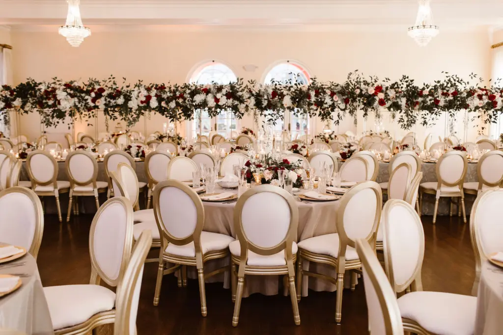 Tall Gold Flower Stand with Cascading Greenery, Burgundy, White, and Blush Rose Centerpiece Inspiration | Gold Chargers and Long Feasting Table | White and Gold Chair Seating for Wedding Reception | South Tampa Venue The Orlo | Planner UNIQUE Weddings + Events