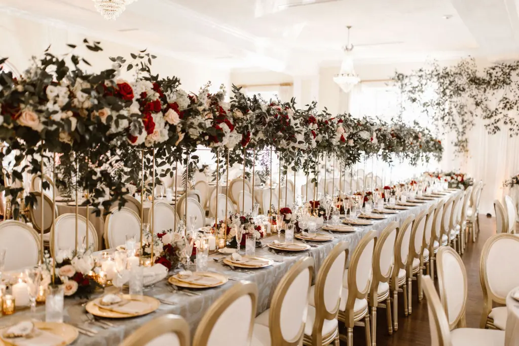 Tall Gold Flower Stand with Cascading Greenery, Burgundy, White, and Blush Rose Centerpiece Inspiration | Gold Chargers and Long Feasting Table | Classic Champagne and Gold Table Setting for Wedding Reception | South Tampa Venue The Orlo | Planner UNIQUE Weddings + Events