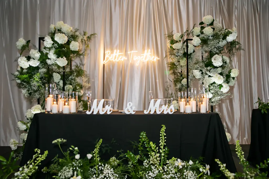 Mr. and Mrs. Sweetheart Table with Pillar Candles in Hurricane Tube Glass | Better Together Neon Sign | Tall Black Metal Flower Stand Backdrop with White Baby's Breath, Hydrangeas, and Greenery | Stock Flower, Snapdragon, and Greenery Floor Flower Arrangement Inspiration