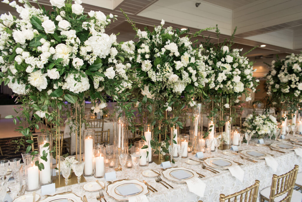 Tall Flower Stand with White Roses, Babies Breath, and Greenery Centerpiece for Wedding Reception Inspiration | Pillar and Taper Candles with Hurricane Glass Tubes | Long Feasting Tables with Gold and White Table Settings | Tampa Bay Rentals A Chair Affair | Planner Parties A La Carte