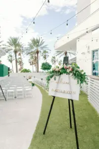 Acrylic Wedding Sign with Gold Writing and Greenery Floral Decor Detailing | White Folding Chairs in Outdoor Tropical Wedding