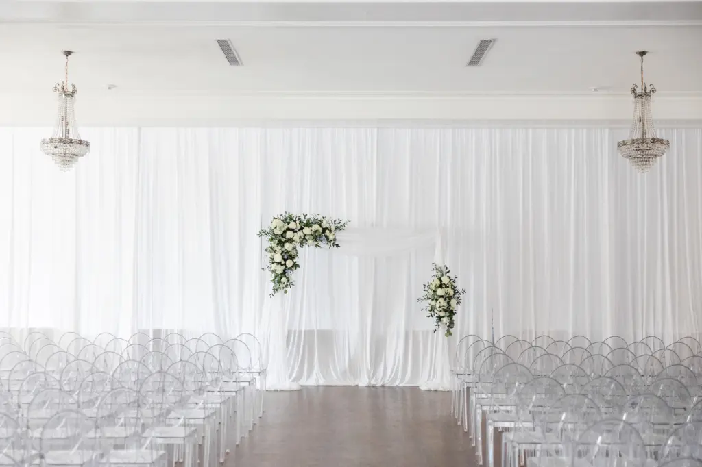 White Drapery and Piping | Asymmetrical Floral Arrangements with White Roses, Blue Flowers, and Greenery Wedding Ceremony Arch Ideas | Ghost Acrylic Chairs | Tampa Bay Kate Ryan Event Rentals | Florist Monarch Events and Design | Planner Kelci Leigh Events