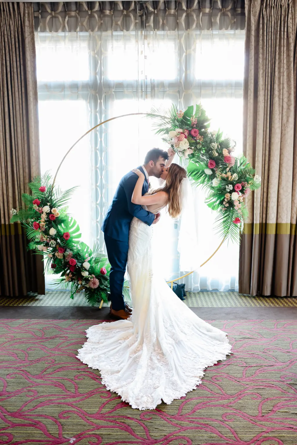 Bride and Groom First Kiss Infront of Gold Circle Arch with Tropical Florals in Indoor Wedding Ceremony | St. Pete Planner Coastal Coordinating | Wedding Photographer Lifelong Photography | Venue The Birchwood