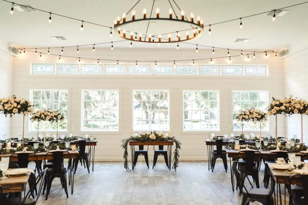 Modern and Rustic Indoor Wedding Reception | Black Metal Chairs | Wooden Tables with Hairpin Legs Seating Inspiration | Tall Flower Stand Centerpieces with White Roses and Greenery | Tampa Bay Florist Monarch Events and Design | Venue Cross Creek Ranch