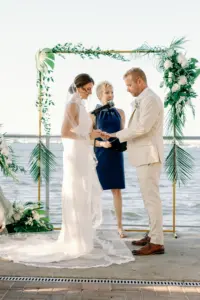 Bride and Groom Exchange Vows in Waterfront Wedding Ceremony in Front of Gold Arch with Greenery Tropical Details Wedding Portrait