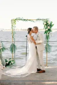Bride and Groom First Kiss in Waterfront Wedding Ceremony in Front of Gold Arch with Greenery Tropical Details Wedding Portrait