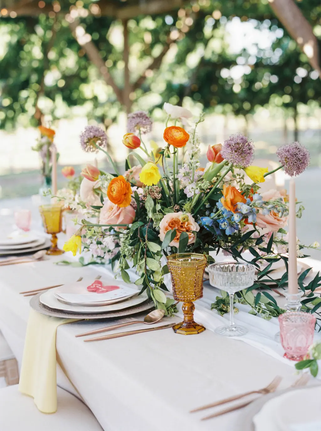 Whimsical Wedding Reception Centerpiece Ideas with Orange Ranunculus, Pink Garden Roses, Purple Allium, Blue Stock Flowers, Tulips, and Yellow Daffodils | Colored Goblets, and Bronze Flatware | Tampa Bay Florist Save The Date Florida