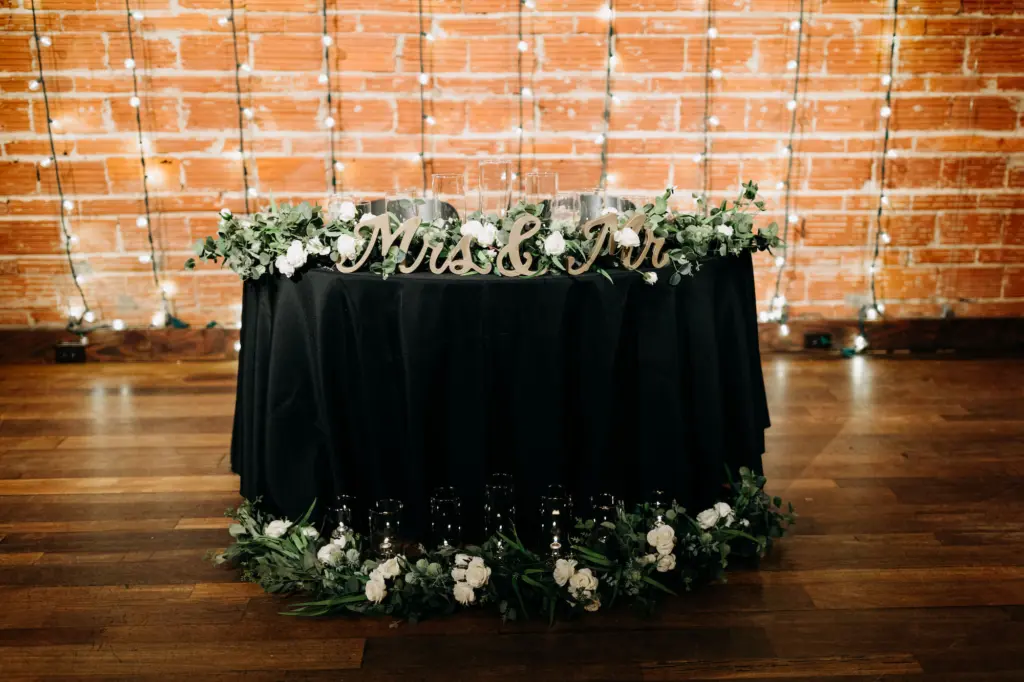 Mrs. and Mr. Sweetheart Table Sign with Black Linen, Greenery and White Roses and String Light Backdrop