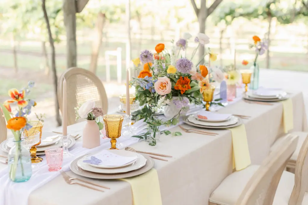 Whimsical Wedding Reception Centerpiece Ideas with Orange Ranunculus, Pink Garden Roses, Purple Allium, Blue Stock Flowers, Tulips, and Yellow Daffodils | White Cheesecloth Table Runners, Yellow Napkins, and Beige Tablecloth | Spring Vineyard Wedding Reception Inspiration | Tampa Bay Florist Save The Date Florida | Planner MDP Events