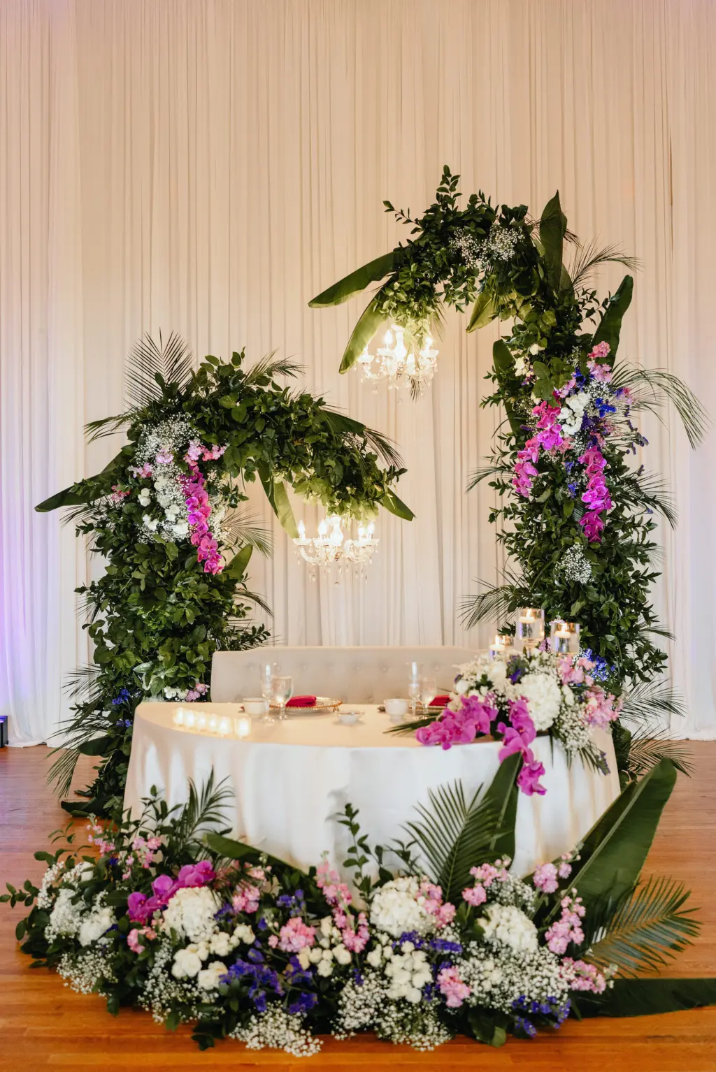 Asian Fusion Floral Sweetheart Table with Unique Wedding Arch of Tropical Leaves and Greenery and Purple Florals Wedding Reception Inspiration