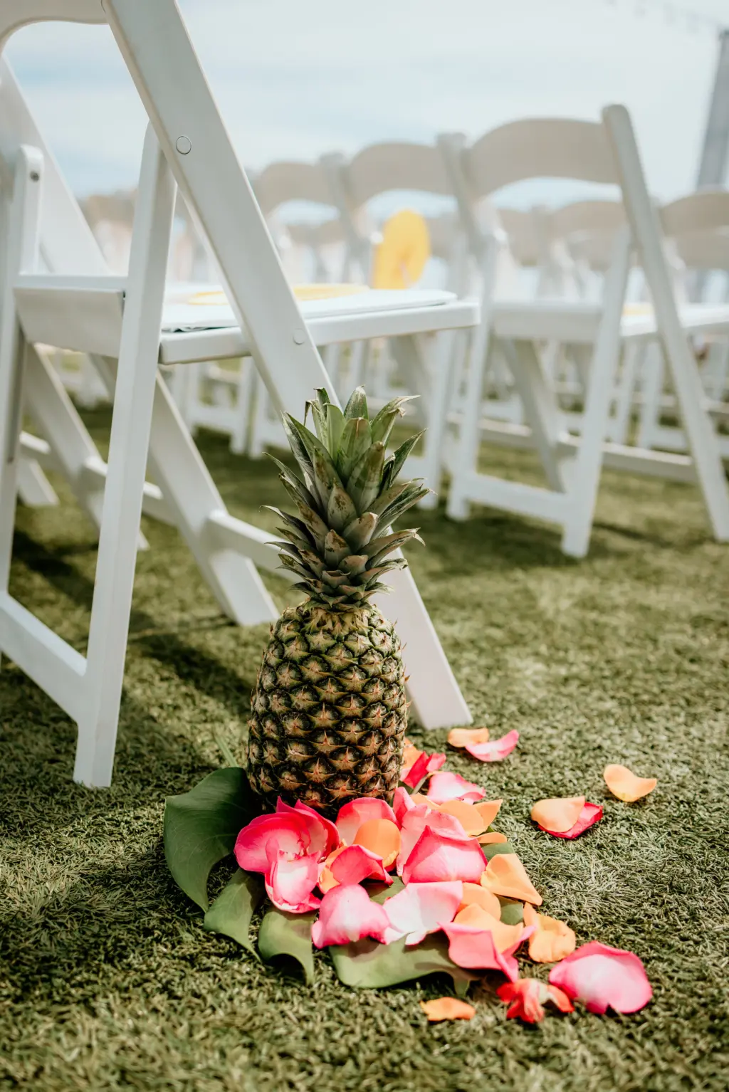 Pineapple and Bright Floral Petal Wedding Aisle Decor with White Folding Chairs | Tropical Outdoor Ceremony Inspiration