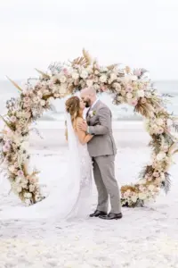 Bride and Groom Wedding Portrait in Beach Ceremony in Front of Circle Boho Wedding Floral Arch Design | Floral Arrangements with Pampas Grass, Tropical Leaves, Dried Florals and Pink Garden Roses Wedding Decor Inspiration