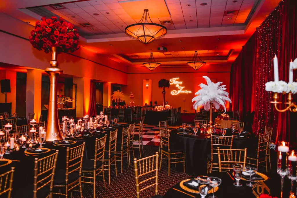 Black Linen Table Cloth with Gold Chiavari Chairs and Chargers | Tall Gold Centerpieces with Candles and White Florals and Feathers Great Gatsby 1920s Tablescape Inspiration | Tampa Rental Company A Chair Affair | Downtown St. Petersburg Florida Event Venue Mahaffey Theatre | Names Couple Monogram Gobo