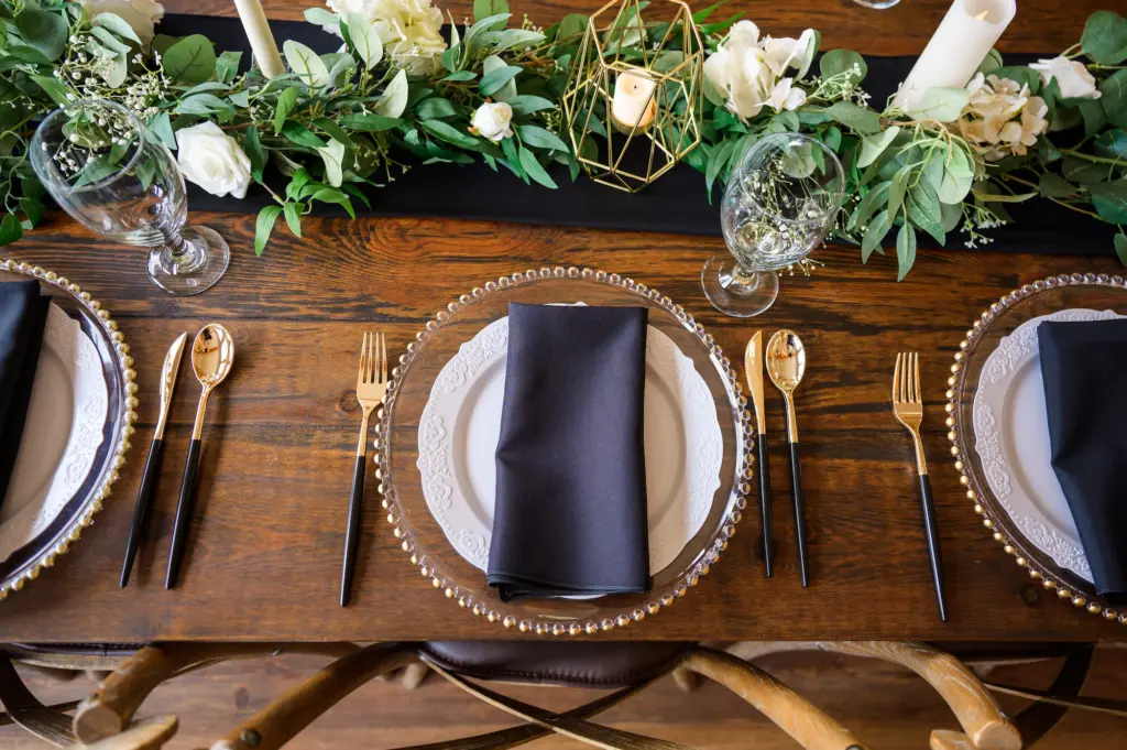 Elegant Southern Wedding Reception Table Setting Inspiration with Black and Gold Flatware, Black Linen, and Beaded Chargers | Wooden Farm Tables with White Flowers and Greenery Garland Centerpiece Ideas