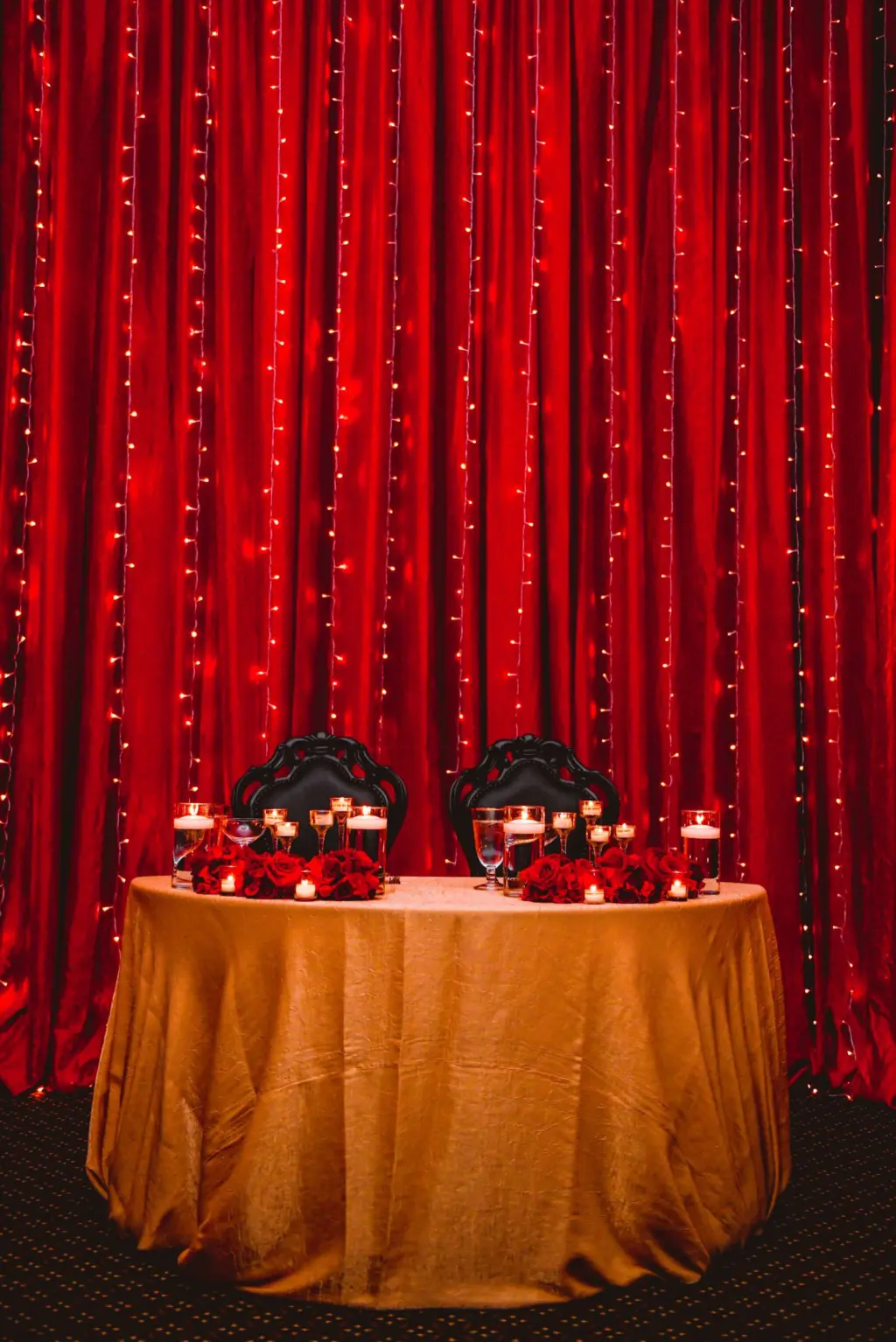 Sweetheart Table with Red Roses and Floating Candles on a Gold Linen with Black Chairs | Tampa Rental Company A Chair Affair | Red Pipe and Drape Backdrop with Twinkle Light Curtain