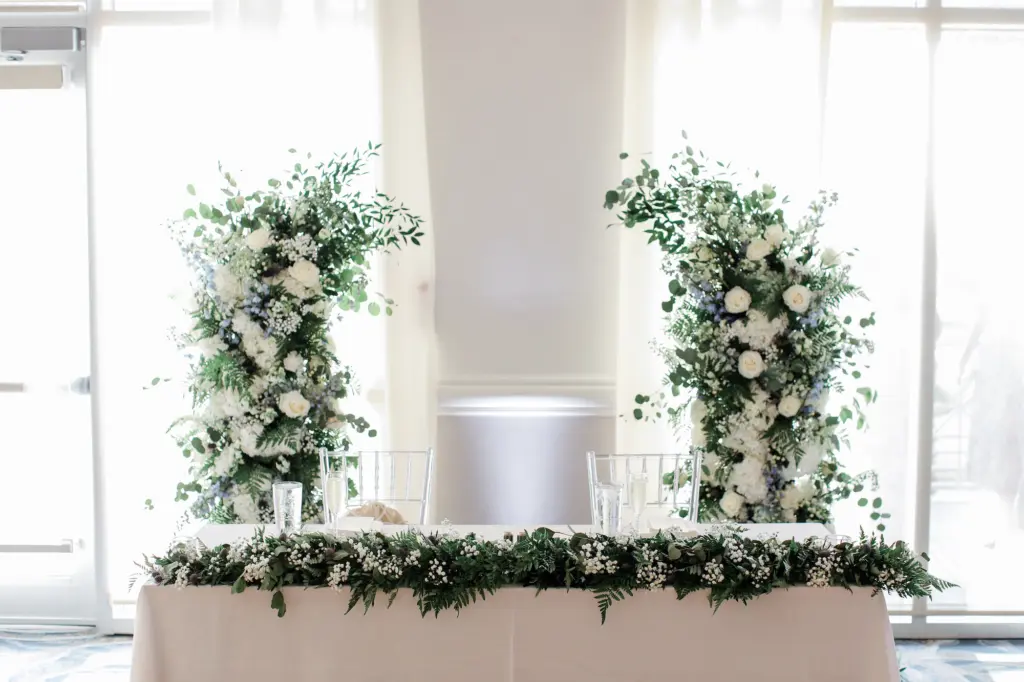 White Roses, Baby's Breath, Hydrangeas, Blue Delphinium, Eucalyptus, and Greenery Floral Backdrop for Sweetheart Table | White and Blue Wedding Reception Decor Ideas | Clear Acrylic Chiavari Chairs | Tampa Bay Event Planner Coastal Coordinating
