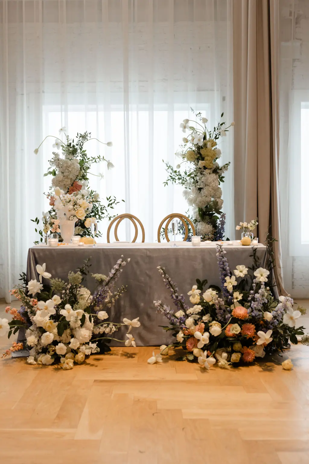 Spring Inspired Sweetheart Table with Gray Linen and White Orchid, Roses, Blue Stock Flowers, Hydrangeas, and Orange Chrysanthemums Wedding Reception Backdrops and Floor Arrangements