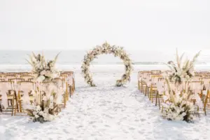 Floral Circle Arch in Boho Beach Wedding Ceremony with Wooden Folding Chairs and Pink Bow Ribbons | Floral Arrangements with Pampas Grass, Hydrangea, White and Champagne Roses and Tropical Greenery Leaves