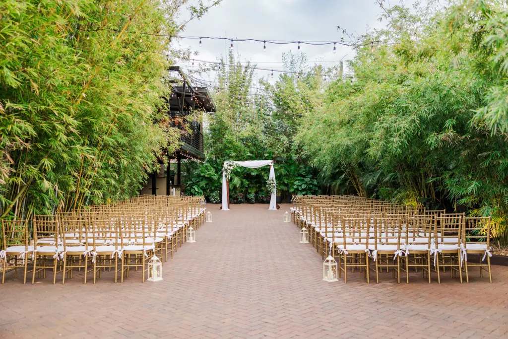 Elegant Gold and White Bamboo Garden Wedding Ceremony Decor Inspiration | Outdoor Ceremony with Gold Chiavari Chairs, Bistro String Lights, and Wood Arch with Fabric Draping | St Pete Event Venue Nova 535 | A Chair Affair Rental Arch Backdrop