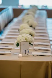 Long Wedding Reception Feasting Table with Camel Mustard Gold Tablecloth | Hydrangea Centerpiece Decor Inspiration | Classic Black and White Table Number Sign Ideas