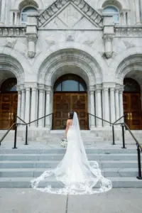 Bride in Front of Downtown Tampa Sacred Heart Catholic Church Wedding Portrait | Lace Deep-V Sheer Lace Bodice Mermaid Maggie Sottero Wedding Dress Ideas | Classic Bridal Hair and Makeup Inspiration with Long Lace Veil | Tampa Videographer J&S Media