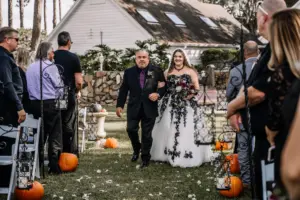 Bride and Father Walking Down Aisle | Gothic Strapless Black Lace, White Tulle, A-Line David's Bridal Wedding Dress Inspiration | Halloween Pumpkin and Black Metal and Crystal Hanging Lantern Aisle Decor Ideas