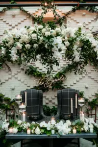 Custom Neon Sign with Lush Greenery Backdrop | Charcoal Plaid Wingback Chairs for Romantic Sweetheart Table Inspiration | White Orchids, Roses, and Greenery Garland and Chandelier Wedding Reception Decor Ideas | Tampa Bay Planner Wilder Mind Events | Downtown Tampa Event Venue Oxford Exchange