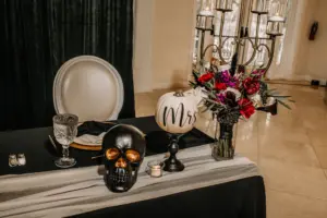 Black and Cream Halloween Wedding Reception Sweetheart Table Inspiration | Pumpkin and Skull Decor Ideas | Red Carnations, Berries, Purple Stock Flowers, White Roses, and Greenery Bouquet Ideas