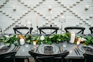Modern Romantic Black and White Conservatory Wedding Reception Inspiration | Floating and Taper Candles, White Roses, and Greenery Garland Centerpiece Decor Ideas | Wooden Black Crossback Chairs | Black-Rimmed Clear Chargers with Modern Gold and Black Flatware