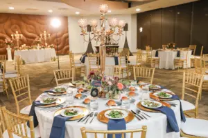 Romantic Pink and Blue Wedding Reception Table Decor Ideas | Gold Chiavari Chairs and Chargers | Hand Lettered Table Numbers | Pink Roses, Spray Roses, Burgundy Chrysanthemums, Blue Thistles, and Greenery Centerpiece Inspiration