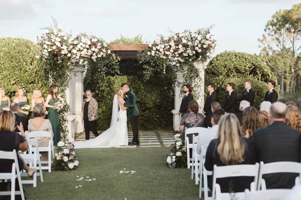 Bride and Groom First Kiss Wedding Portrait | Luxurious Emerald Green Outdoor Wedding Ceremony on the Event Lawn Ideas | Pergola with White Roses, Purple Chrysanthemums, and Cascading Greenery Decor Inspiration | Tampa Bay Venue The Concession Golf Club | Florist Bruce Wayne Florals