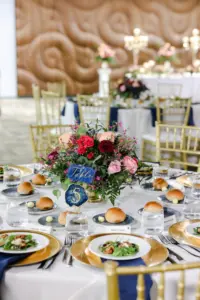 Romantic Pink and Blue Wedding Reception Table Decor Ideas | Gold Chiavari Chairs and Chargers | Hand Painted Table Numbers | Pink Roses, Spray Roses, Burgundy Chrysanthemums, Blue Thistles, and Greenery Centerpiece Inspiration