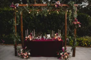 Whimsical Boho Wedding Reception Sweetheart Table Under Pergola with Greenery Decor Inspiration | Pink and Burgundy Roses, and Stock Flower Arrangement Ideas