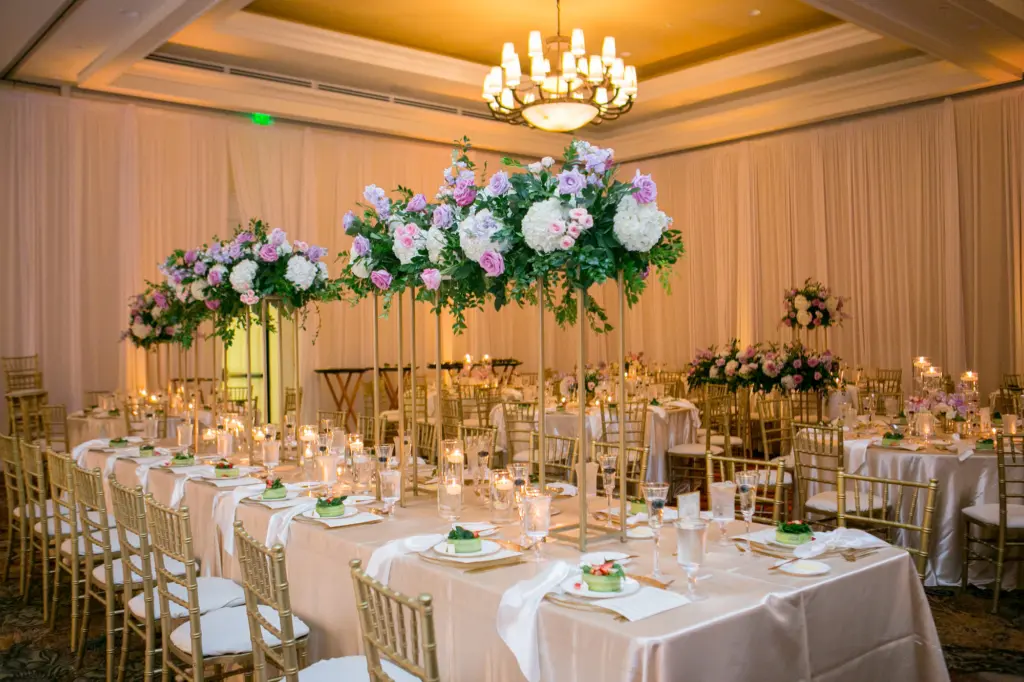 Elegant Wedding Reception Long Feasting Table Decor Inspiration | Satin Ivory Tablecloths | Gold Flatware, Chargers, Chiavari Chairs | Tall Flower Stand with Pink and Purple Roses, Greenery, and White Hydrangeas Centerpiece Ideas | Tampa Bay Kate Ryan Event Rentals | Clearwater Beach Event Venue Sandpearl Resort Hotel Ballroom