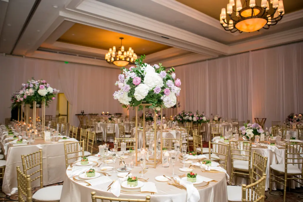 Elegant Wedding Reception Table Decor Inspiration | Satin Ivory Tablecloths | Gold Flatware, Chargers, Chiavari Chairs | Tall Flower Stand with Pink and Purple Roses, Greenery, and White Hydrangeas Centerpiece Ideas | Tampa Bay Kate Ryan Event Rentals | Clearwater Beach Event Venue Sandpearl Resort Hotel Ballroom