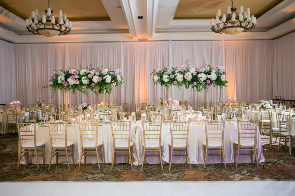 Elegant Wedding Reception Long Feasting Table Decor Inspiration | Satin Ivory Tablecloths | Gold Flatware, Chargers, Chiavari Chairs | Tall Flower Stand with Pink and Purple Roses, Greenery, and White Hydrangeas Centerpiece Ideas | Tampa Bay Kate Ryan Event Rentals | Clearwater Beach Event Venue Sandpearl Resort Hotel Ballroom