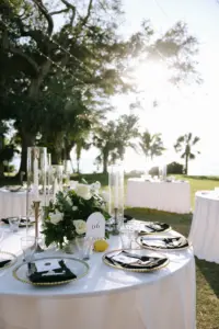 Elegant Black and White Outdoor Wedding Reception Table Setting Ideas | White Roses, Greenery, Taper Candles, Hurricane Glass Cylinders, and Lemon Italian Centerpiece Inspiration | Round Tables with White Linens and Black Napkins on Gold Beaded Glass Charger Plates