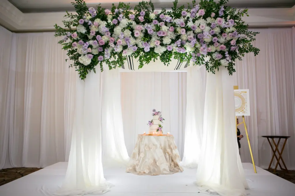 Whimsical Chuppah Over Cake Table Inspiration | White Round Four-tiered Buttercream Wedding Cake with Purple and Pink Rose Accents | Jewish Chuppah Decor with White Drapery, Pink and Purple Roses, Greenery and White Hydrangea Flower Arrangement