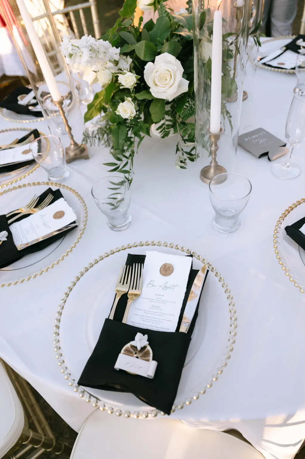 Glamorous Gold, White, and Black Wedding Reception Table Setting Decor Ideas | Gold Beaded Glass Chargers, Black Napkin Linen, White Rose, Greenery and Taper Candle Centerpiece Inspiration | Place Setting Menu Card with Wax Seal