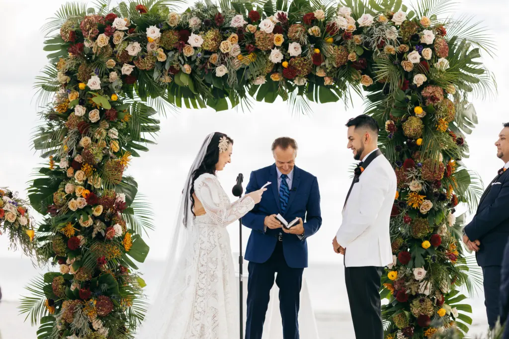 Bride and Groom Vow Exchange | Tropical Fall Moody Black Tie Beach Wedding Ceremony Arch Inspiration | White, Yellow, and Blush Roses, Burgundy Chrysanthemums, Monstera Leaves, and Palm Leaves