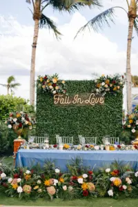 Fall in Love Neon Sign With Grass Boxwood Hedge Wall Backdrop Inspiration | Flower Arrangement for Wedding Reception Sweetheart Table Decor Ideas | Yellow Mums, White Roses, Burgundy Chrysanthemums, Palm Fronds. and Eucalyptus
