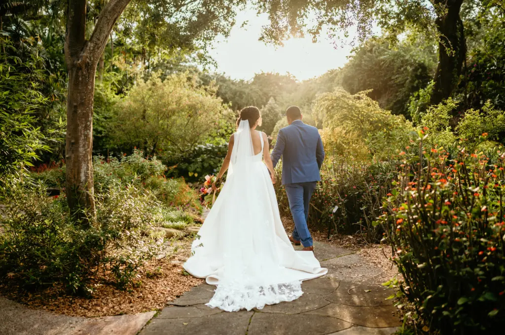 Bride and Groom Sunset Photo Walking Through the Garden | White Lace A-Line Wedding Dress Ideas | Dusty Blue Groom Suit Inspiration | Downtown St. Pete Event Outdoor Venue Sunken Gardens