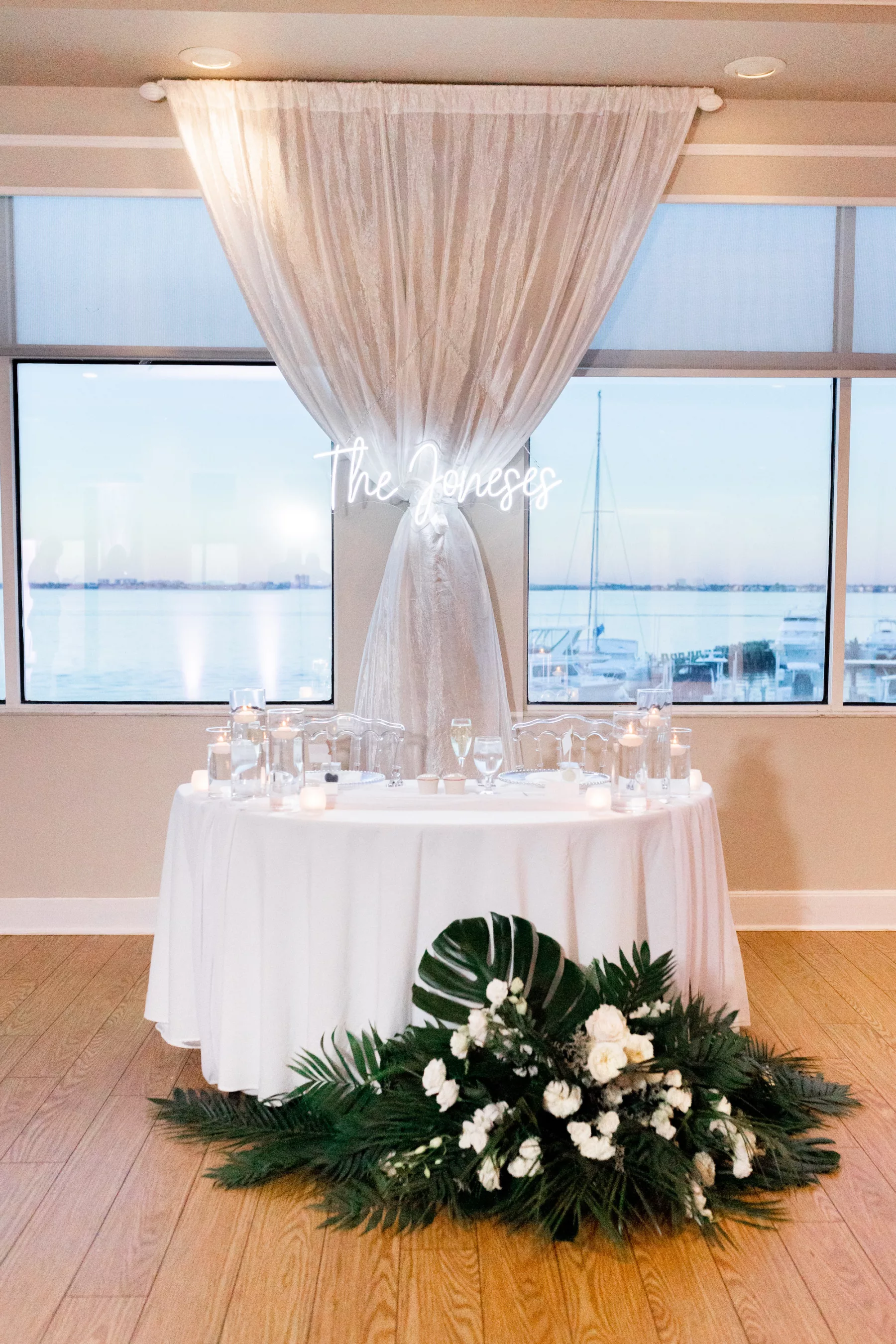 Modern Tropical Indoor Wedding Reception Sweetheart Table Inspiration | Custom Neon Sign, White Linen, and Floral Arrangement with Monstera Leaves, Palm Leaves, White Roses, and Snap Dragons