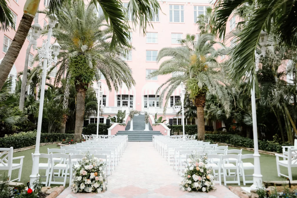 Elegant White Outdoor Courtyard Wedding Ceremony Inspiration | Garden Folding Chairs | White Roses, Pink Carnations, Orange Anemone, and Greenery Aisle Decor Ideas | St Pete Event Venue The Don Cesar | Photographer Lifelong Photography Studio