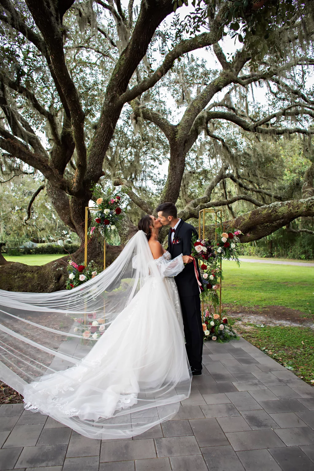 Bride and Groom First Kiss Wedding Portrait | Gold Wedding Ceremony Tree Arch with Red Roses, Blue Thistle, Pink Roses, Peach Chrysanthemums, White Bacopa, and Greenery Decor Ideas | Tampa Bay Florist Save The Date Florida | Event Venue Legacy Lane Weddings | Brooksville Photographer Limelight Photography | Videographer Priceless Studios