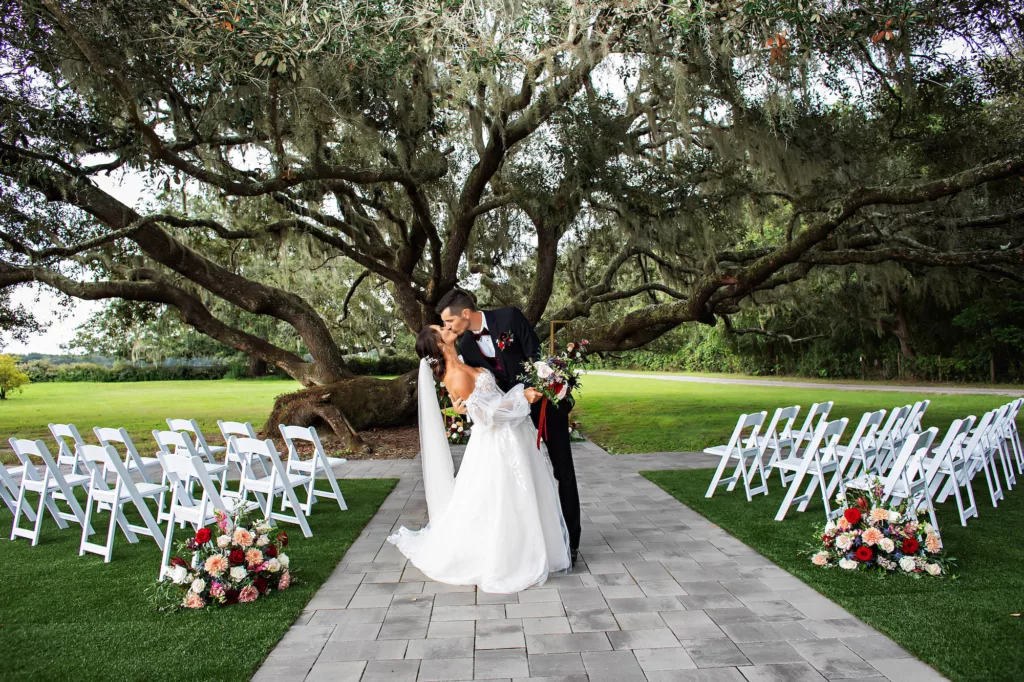 Bride and Groom Just Married Wedding Portrait | Fall Wedding Ceremony Tree Ideas | Folding Garden Chairs | Red Roses, Blue Thistle, Pink Roses, Peach Chrysanthemums, White Bacopa, and Greenery Aisle Decor Ideas | Tampa Bay Florist Save The Date Florida | Event Venue Legacy Lane Weddings | Brooksville Photographer Limelight Photography | Videographer Priceless Studios
